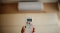 Hemat listrik AC - remote suhu Tangan memegang remote AC dengan unit AC split putih di dinding sebagai latar