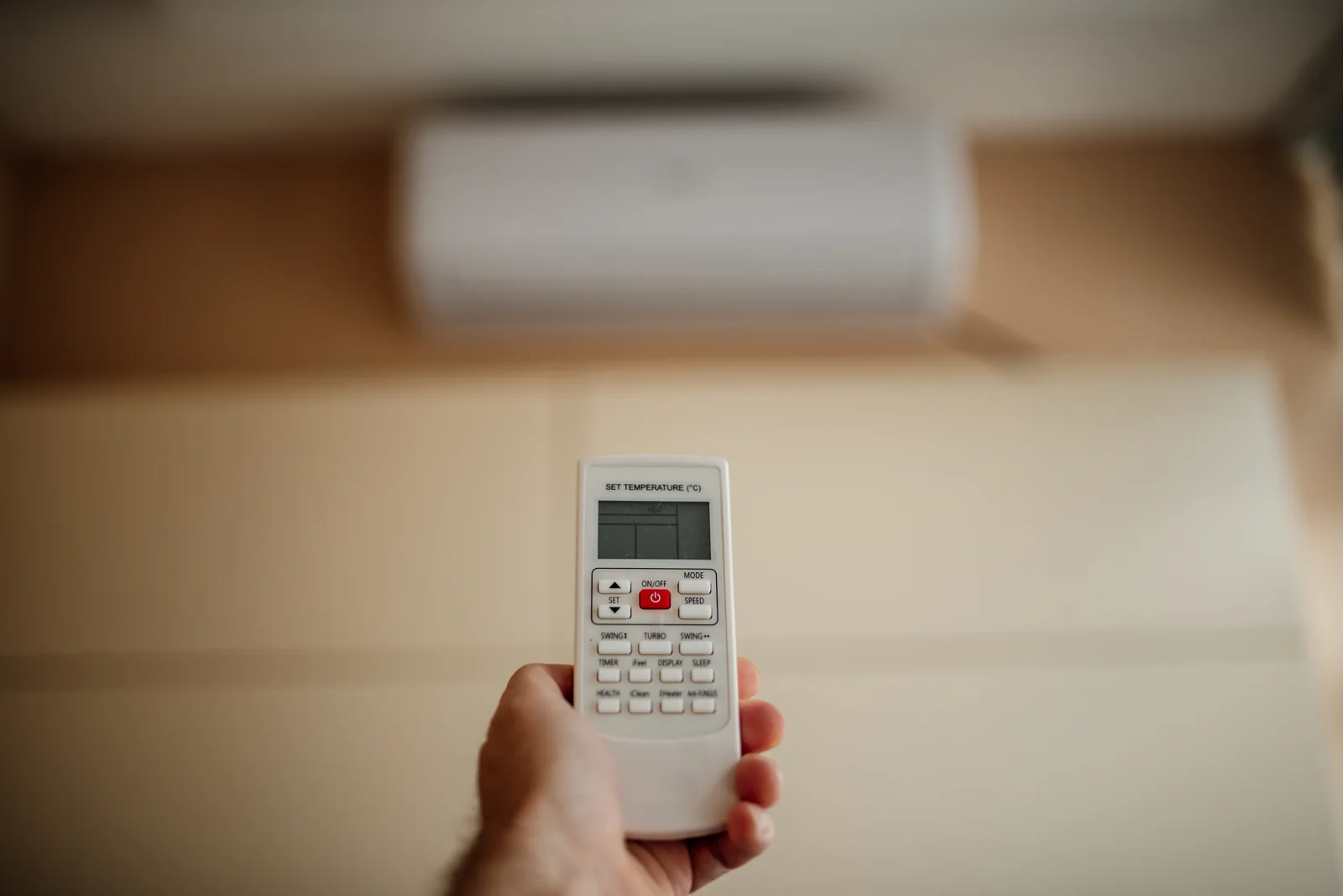 Tangan memegang remote AC dengan unit AC split putih di dinding sebagai latar
