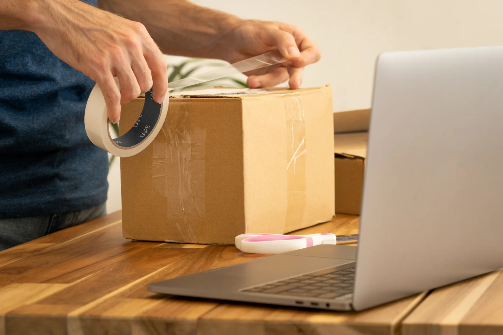 Tangan mengemas paket dengan lakban di depan laptop terbuka untuk pengiriman pesanan online