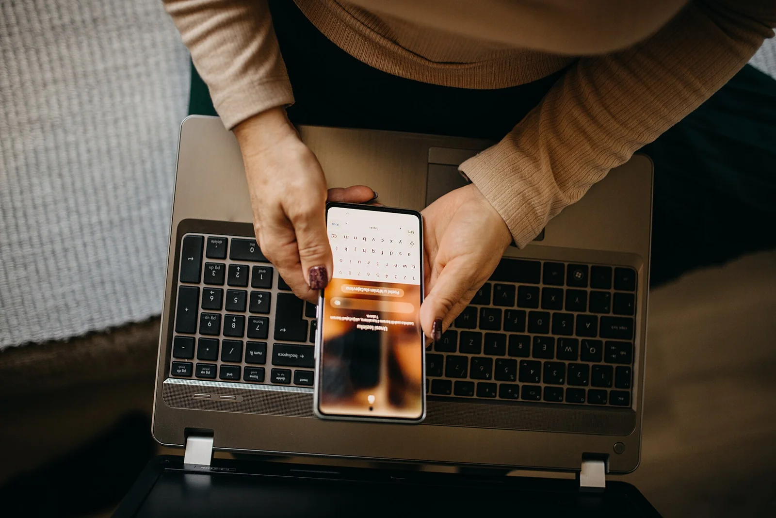 Seorang perempuan memegang ponsel di atas laptop terbuka, dilihat dari atas