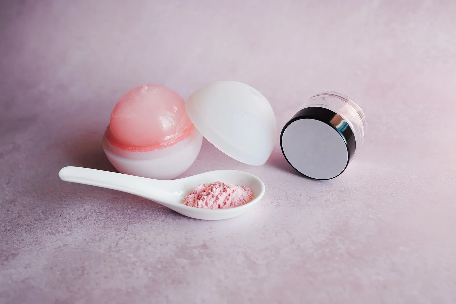 Beberapa jar produk skincare dan sendok berisi bubuk merah muda di atas latar merah muda