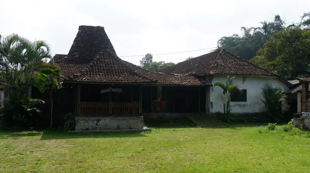 Rumah joglo beratap tinggi dikelilingi pepohonan dan halaman rumput