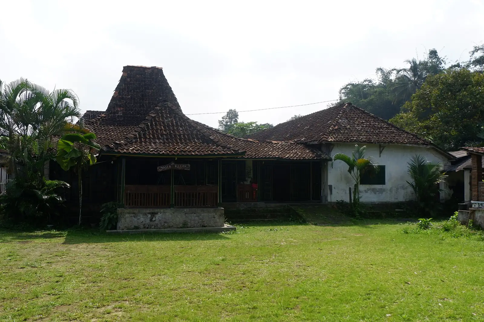 Rumah joglo beratap tinggi dikelilingi pepohonan dan halaman rumput