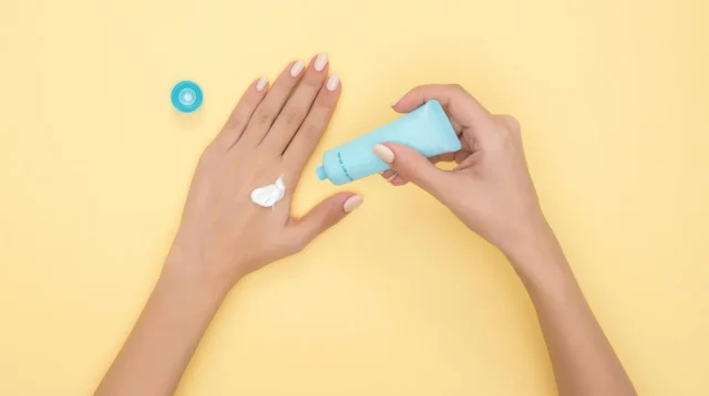 Tangan perempuan mengoles krim pelembap dari tube biru di atas latar kuning