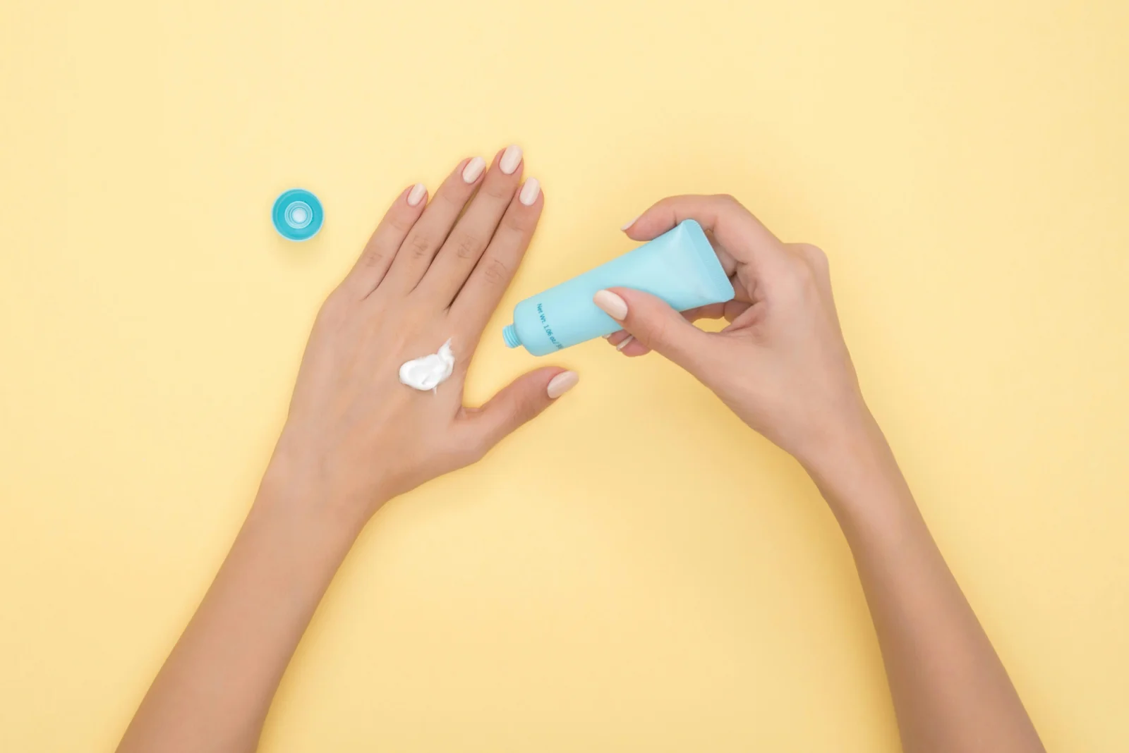 Tangan perempuan mengoles krim pelembap dari tube biru di atas latar kuning