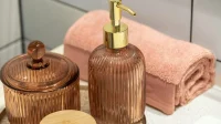 Botol pump kaca bergaris dan toples berwarna peach di dekat handuk tergulung di wastafel