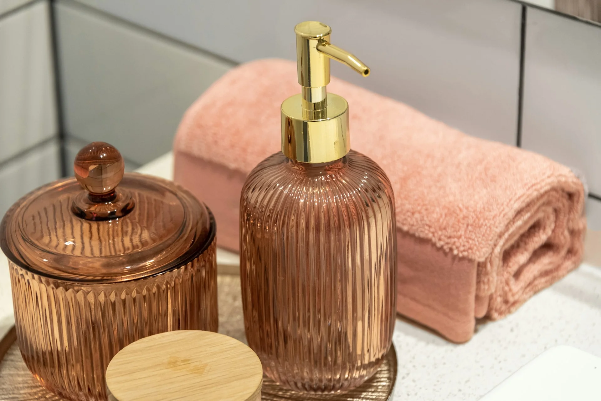 Botol pump kaca bergaris dan toples berwarna peach di dekat handuk tergulung di wastafel