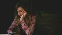 Perempuan bertopang dagu menatap ponsel di meja, ilustrasi cara mengatasi hp lemot