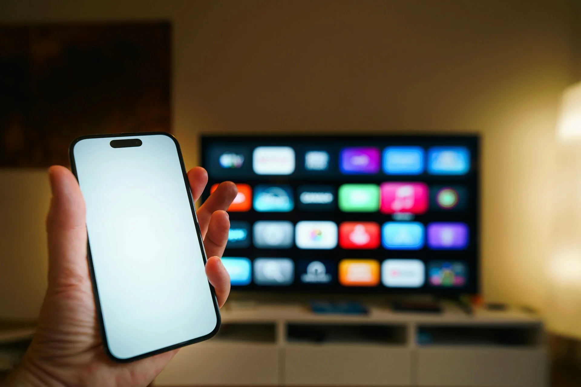Tangan memegang ponsel di depan smart TV yang menampilkan deretan aplikasi di ruang keluarga
