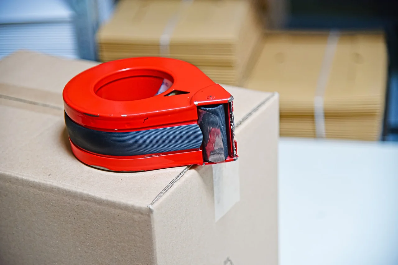 Dispenser lakban merah di atas kardus dengan tumpukan kardus lipat di belakangnya