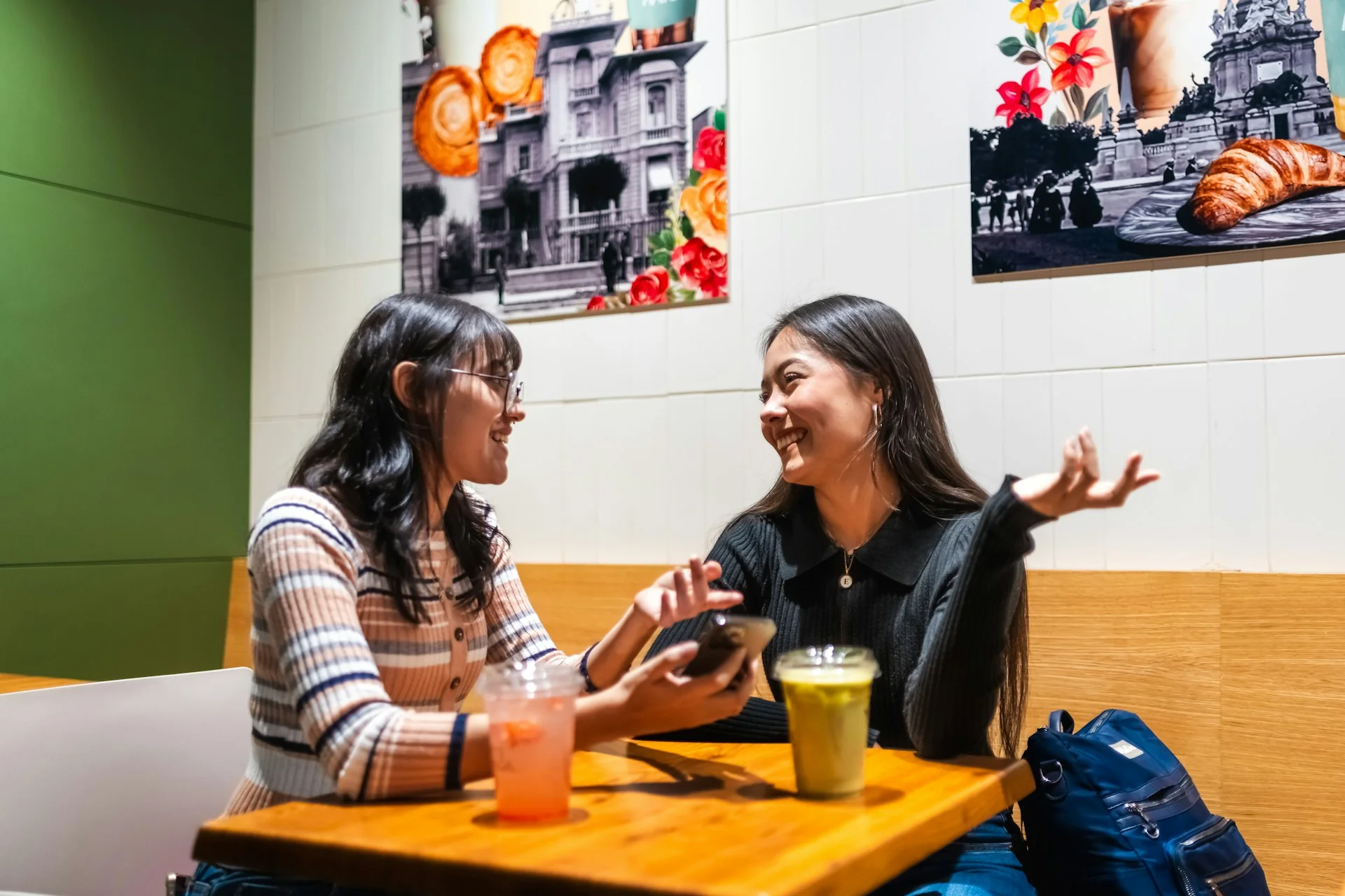 Dua perempuan muda duduk di meja kafe, satu bercerita sambil menggerakkan tangan dan satu lagi menyimak sambil tersenyum