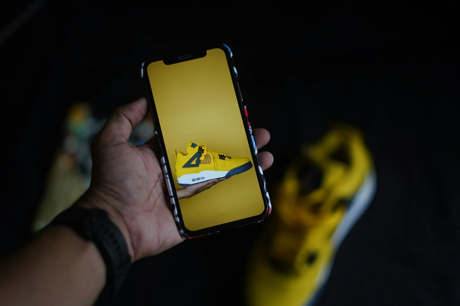 Tangan memegang HP yang menampilkan foto sepatu kuning berlatar polos, dengan sepatu aslinya buram di belakang