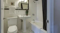 Kamar mandi bersih dengan keramik putih, wastafel, kloset, dan bak mandi bertirai gelap