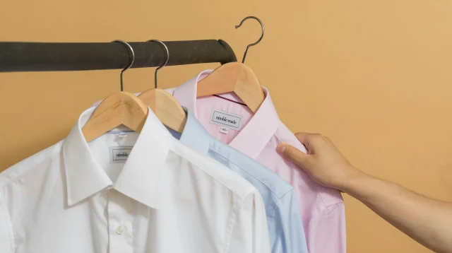 Kemeja putih, biru muda, dan merah muda tergantung di hanger kayu pada rak baju