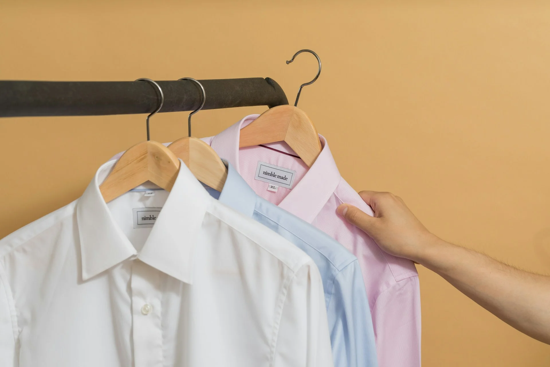 Kemeja putih, biru muda, dan merah muda tergantung di hanger kayu pada rak baju
