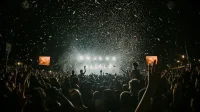Kerumunan penonton konser mengangkat tangan di depan panggung bercahaya terang dengan hujan confetti