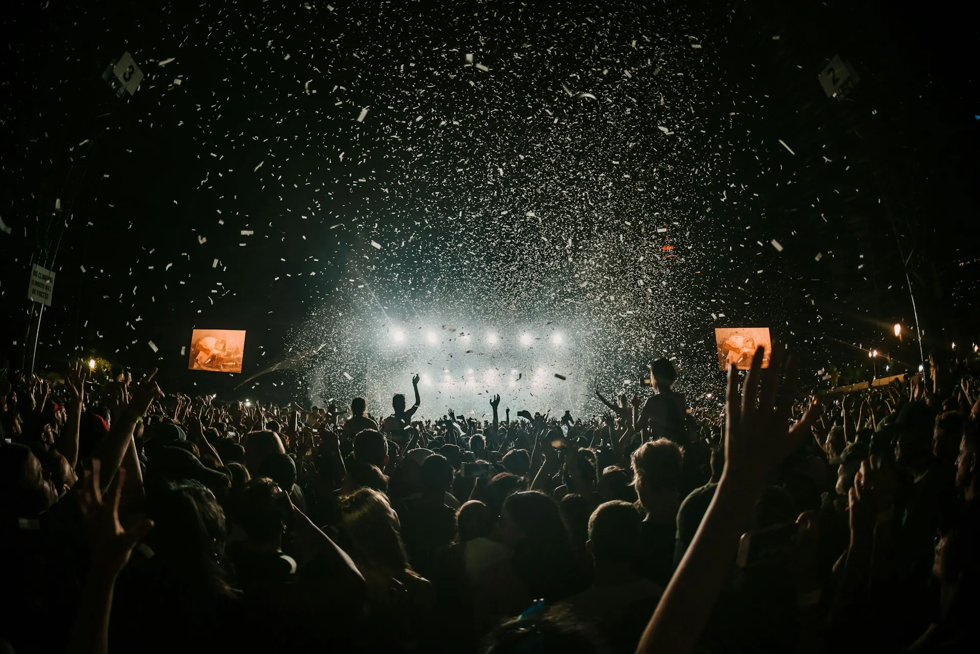 Kerumunan penonton konser mengangkat tangan di depan panggung bercahaya terang dengan hujan confetti