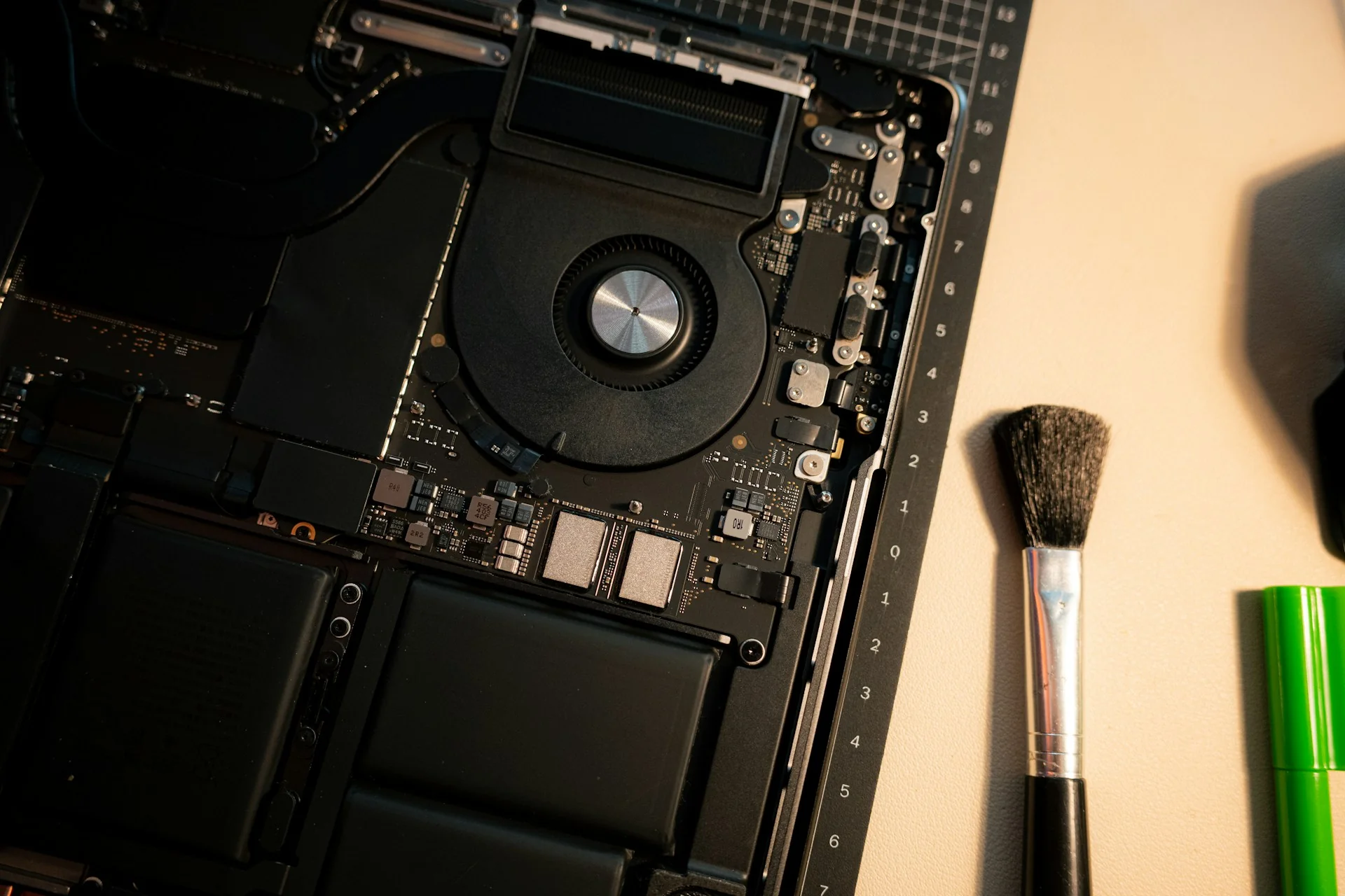 Bagian dalam laptop yang dibuka memperlihatkan kipas pendingin dan papan sirkuit, dengan kuas kecil di sampingnya