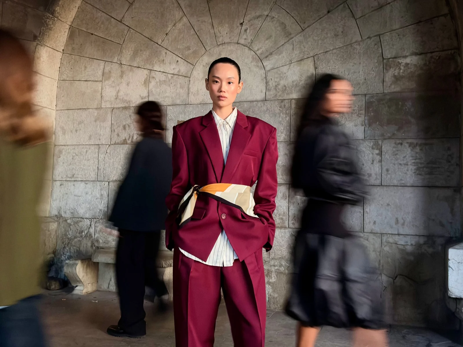 Contoh foto iPhone 18 Pro: model berjas merah marun diam di lorong batu sementara orang di sekitarnya tampak kabur