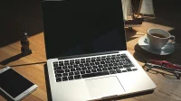 Laptop perak terbuka di atas meja kayu dengan secangkir kopi, kacamata, dan ponsel di sampingnya
