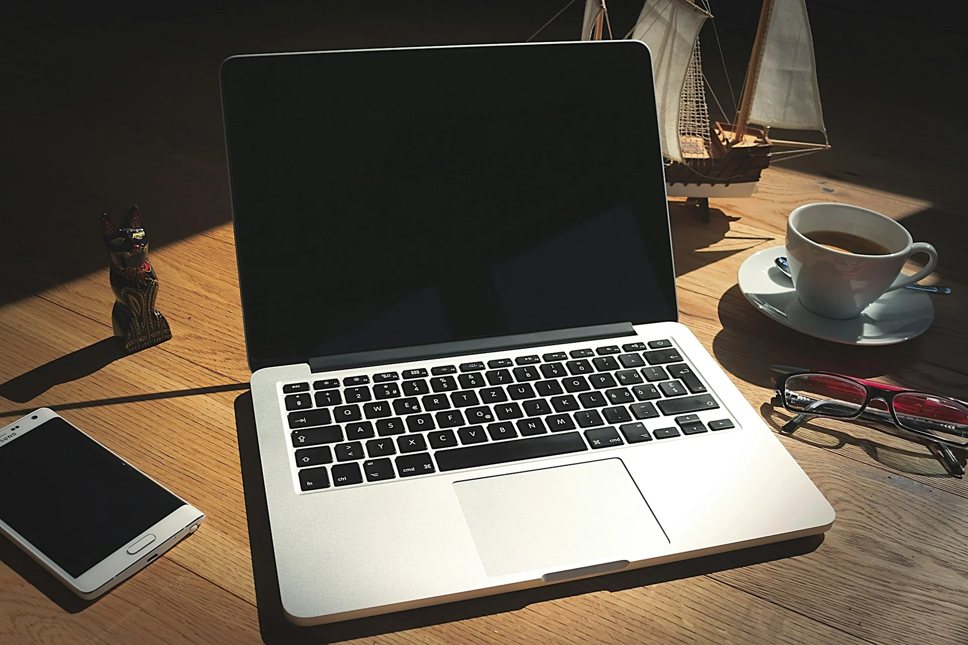 Laptop perak terbuka di atas meja kayu dengan secangkir kopi, kacamata, dan ponsel di sampingnya