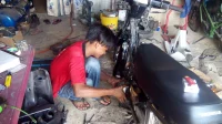 Mekanik berbaju merah berjongkok memeriksa sepeda motor Honda di bengkel