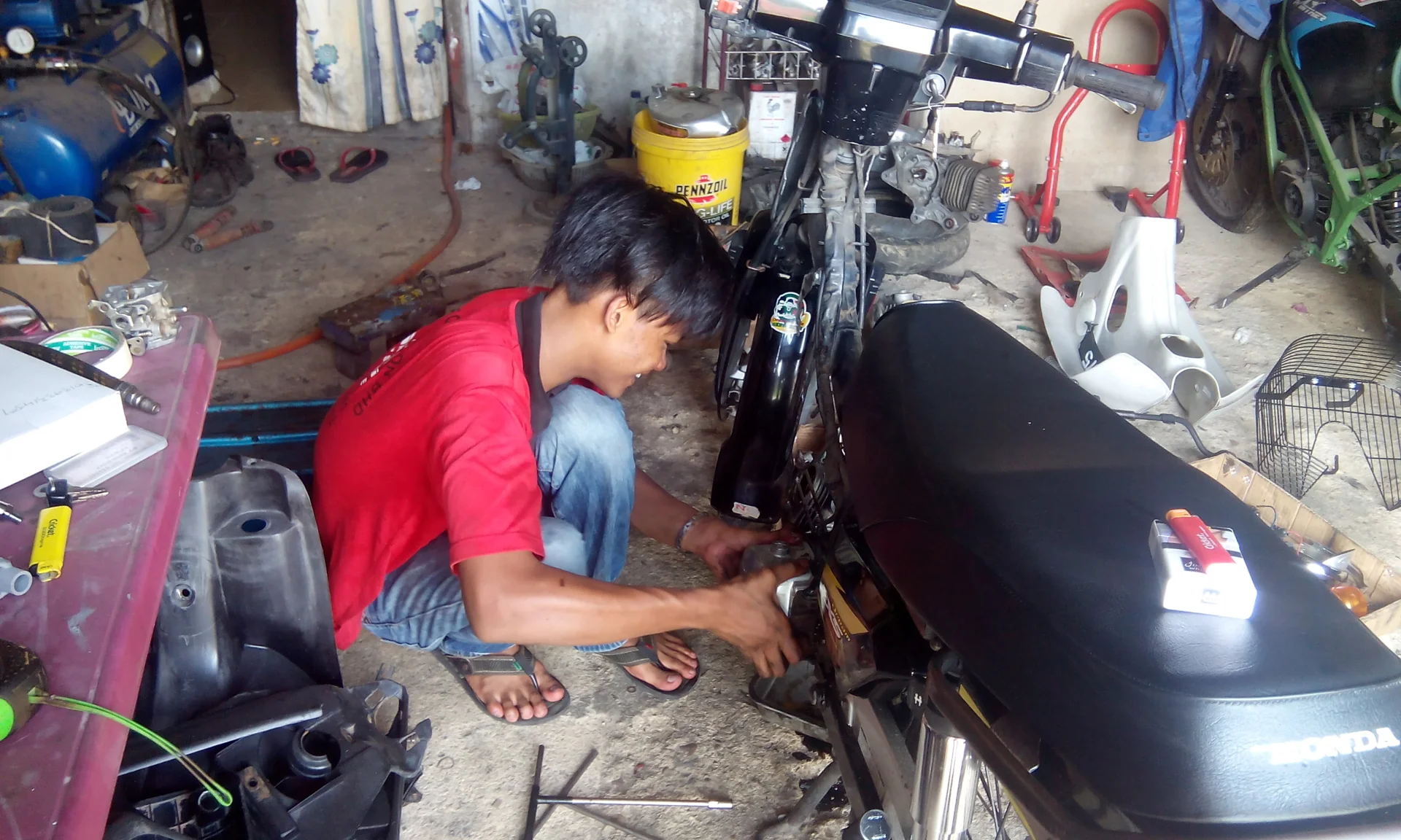 Mekanik berbaju merah berjongkok memeriksa sepeda motor Honda di bengkel