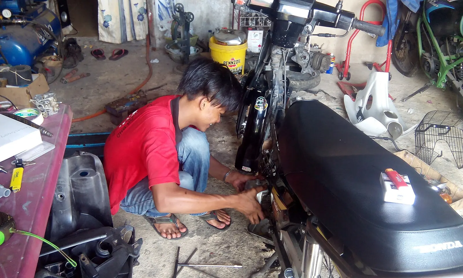 Mekanik berkaos merah jongkok memperbaiki bagian bawah sepeda motor Honda di bengkel kecil