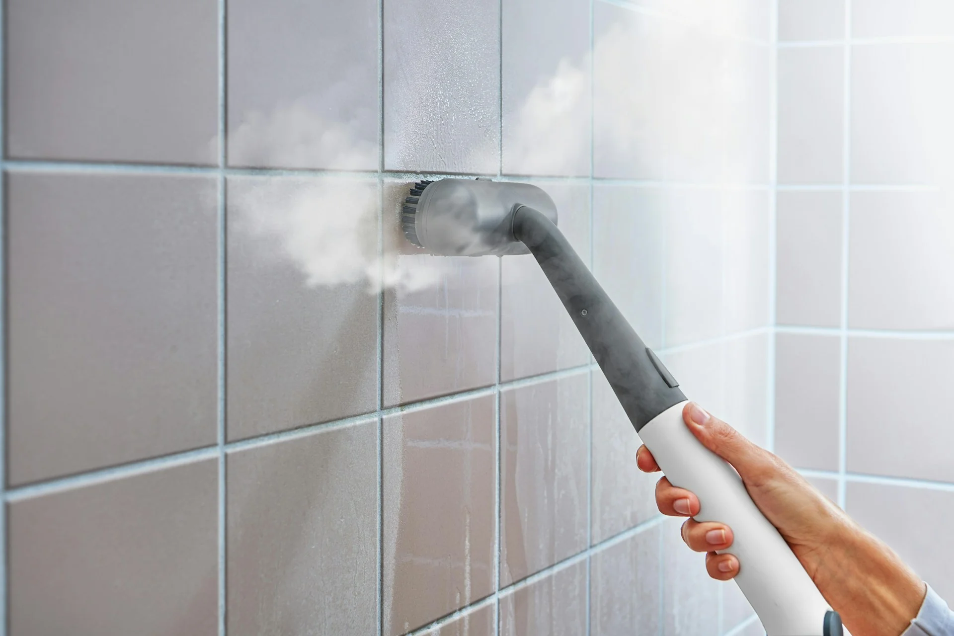 Tangan membersihkan nat keramik dinding kamar mandi dengan sikat steam cleaner yang mengeluarkan uap