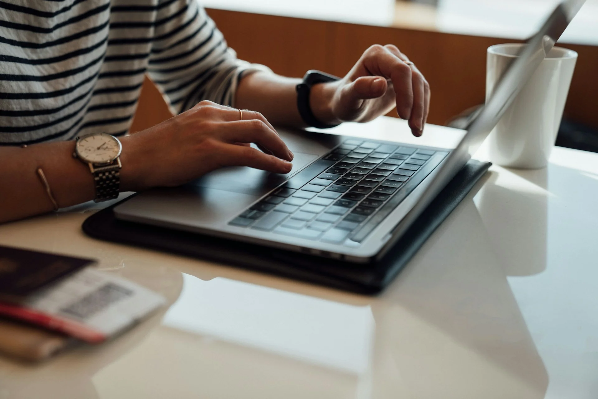 Tangan seseorang mengetik di laptop di atas meja putih dengan cangkir di sampingnya