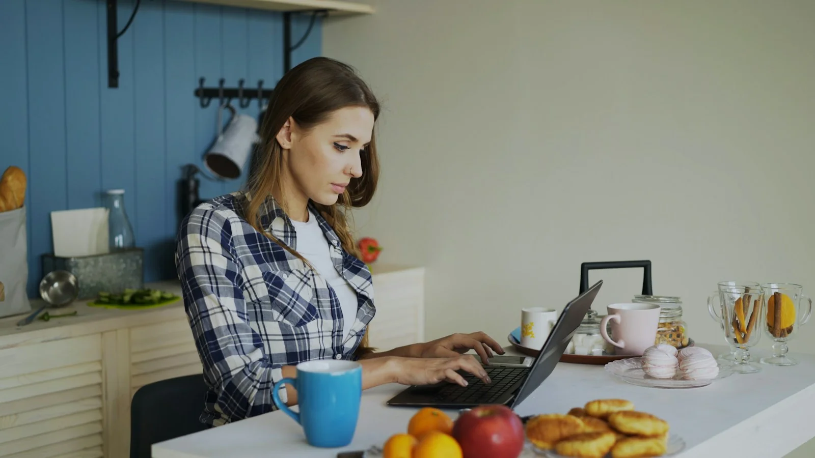 Perempuan mengetik di laptop di meja dapur rumah dengan kue di dekatnya, gambaran mengurus PIRT online