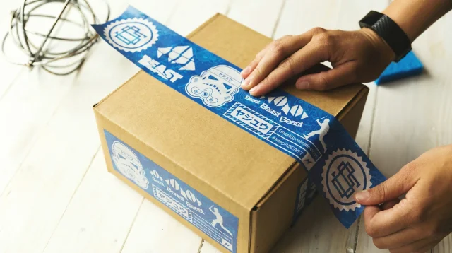 Tangan menyegel kardus paket stok barang jualan online dengan lakban biru di atas meja kayu putih