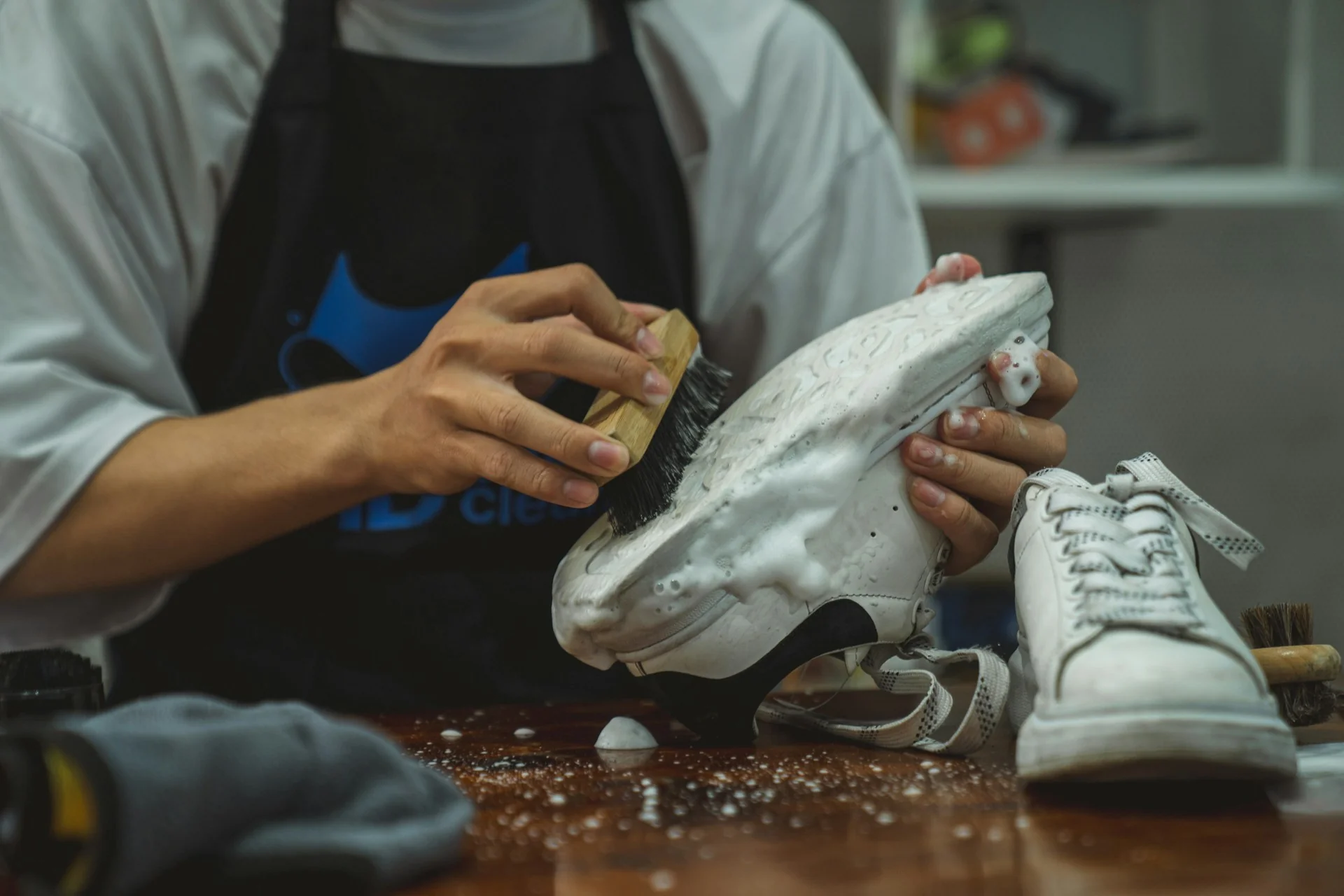Tangan menyikat sneaker kulit putih yang berbusa dengan sikat sepatu di atas meja kayu