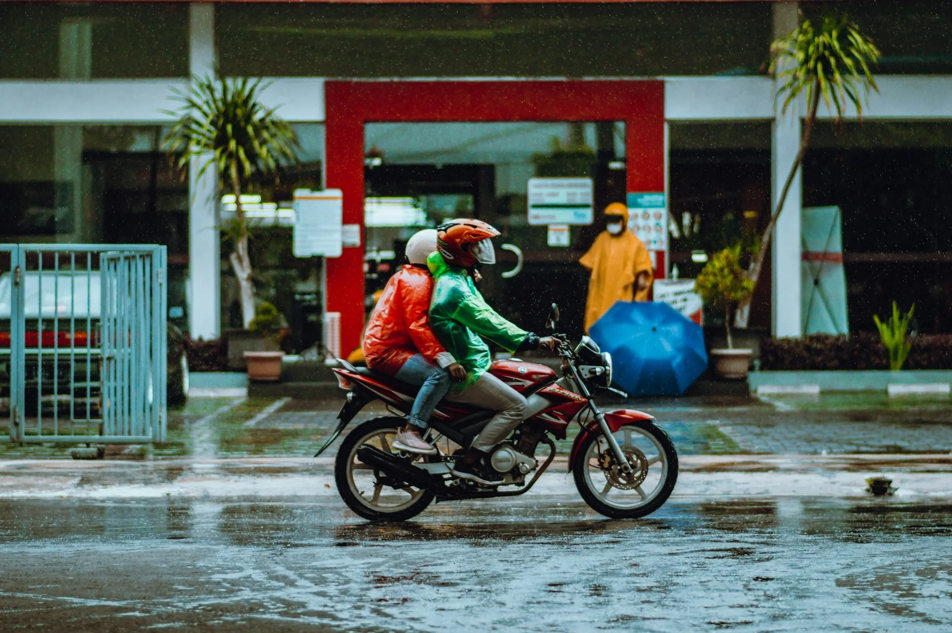 Dua pengendara motor berjaket dan jas hujan melintas di jalan basah saat hujan
