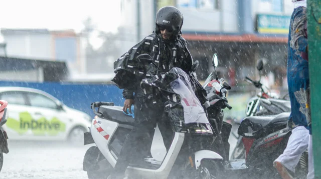 Motor listrik kena hujan deras, pengendara berhelm dan berjas hujan duduk di atas skuter listrik putih