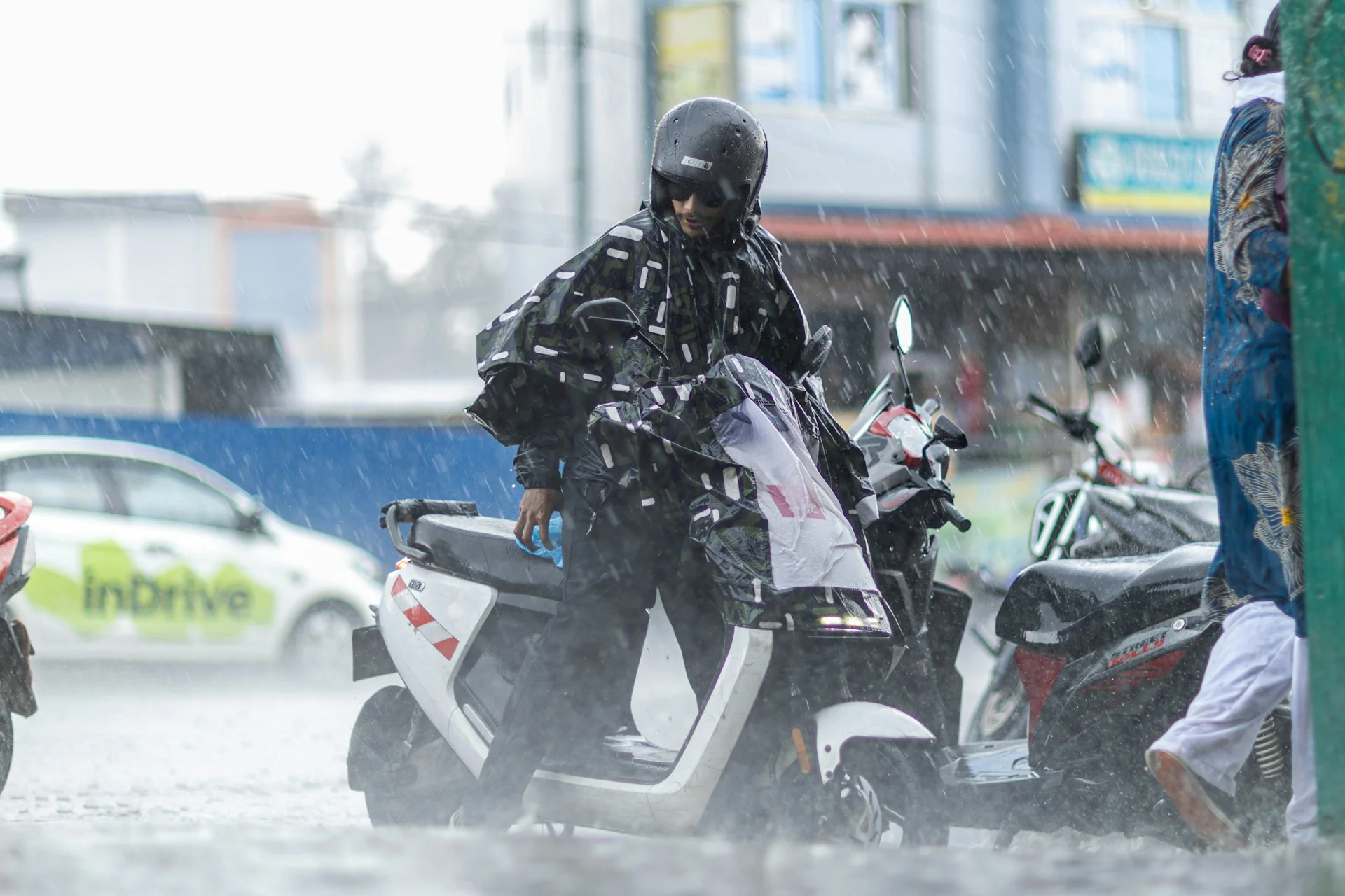 Motor listrik kena hujan deras, pengendara berhelm dan berjas hujan duduk di atas skuter listrik putih