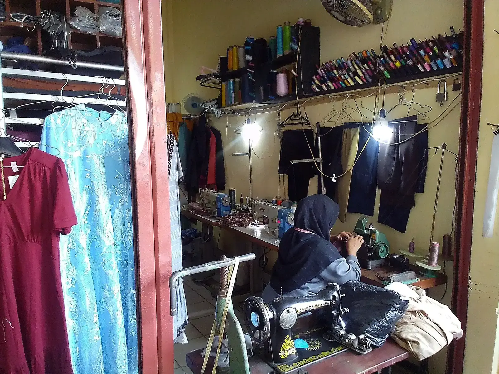 Penjahit berkerudung bekerja di depan mesin jahit di kios jahit kecil dengan gulungan benang dan pakaian tergantung