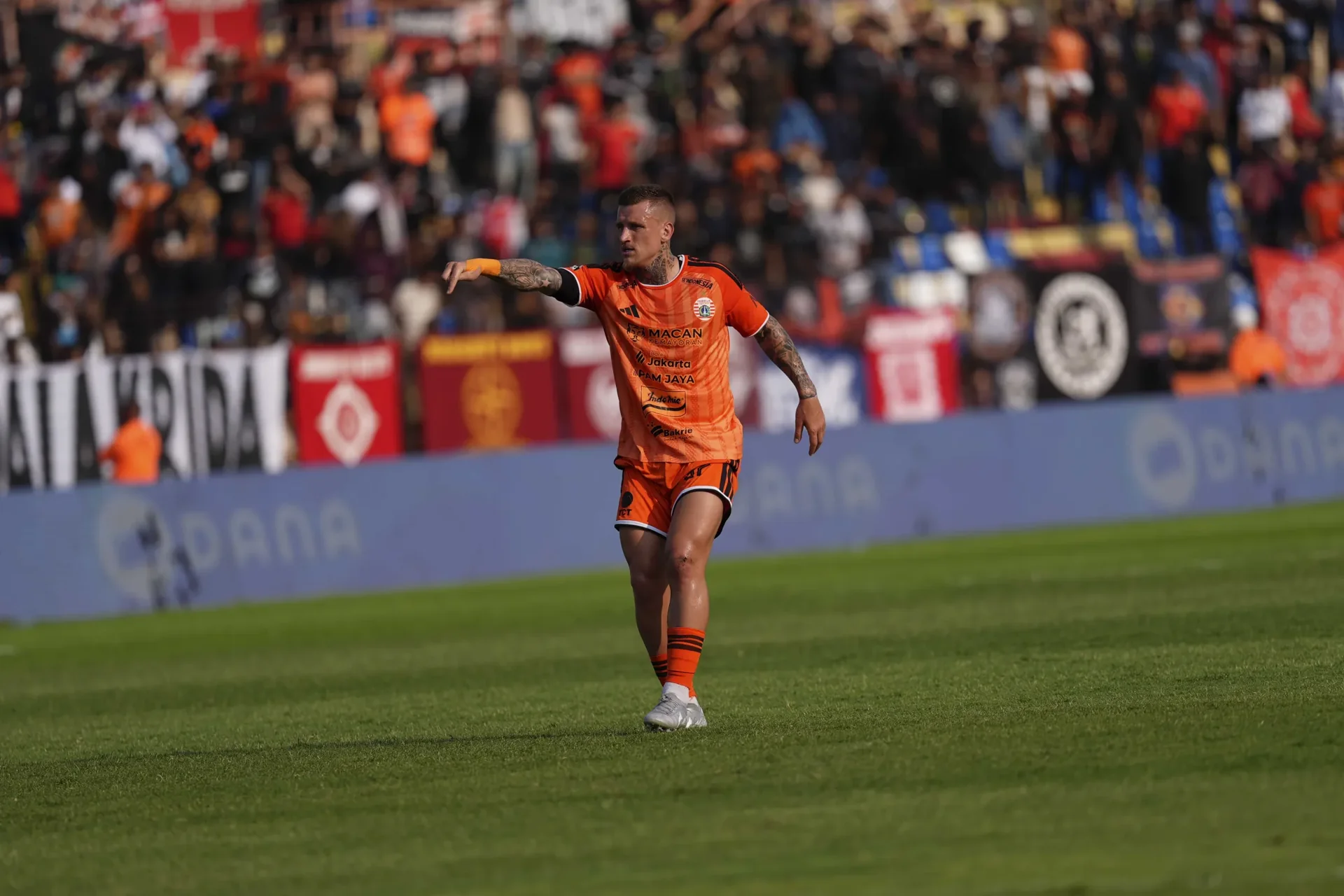 Pemain Persija Jakarta berseragam oranye memberi arahan di lapangan dengan latar suporter