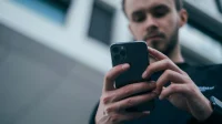 Pria berkaus hitam memegang smartphone dengan dua tangan dan menatap layarnya