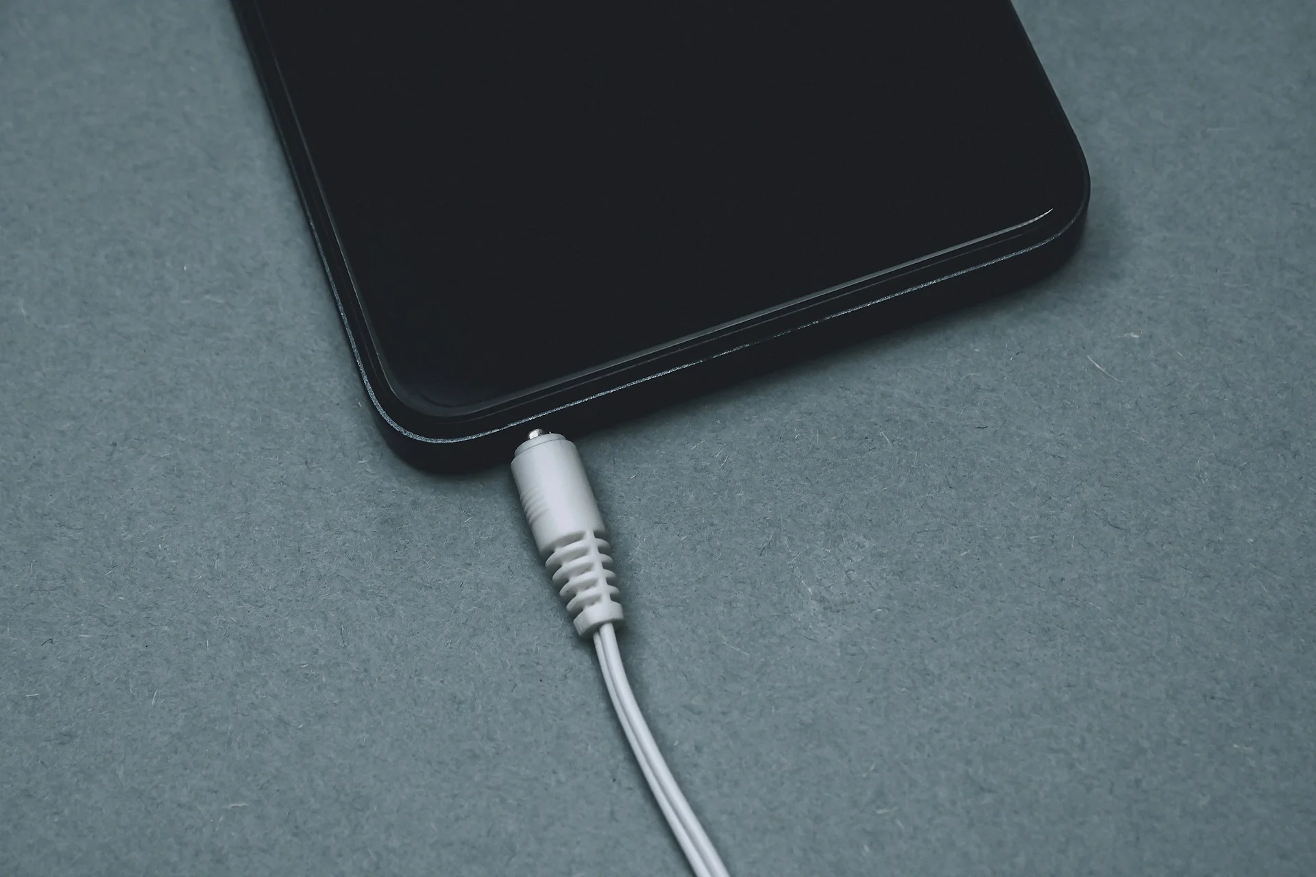 Smartphone hitam tergeletak di permukaan abu-abu dengan kabel putih tersambung ke port bawah