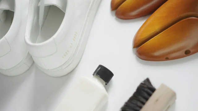 Sepasang sneaker putih di samping sikat sepatu, botol cairan pembersih, dan shoe tree kayu