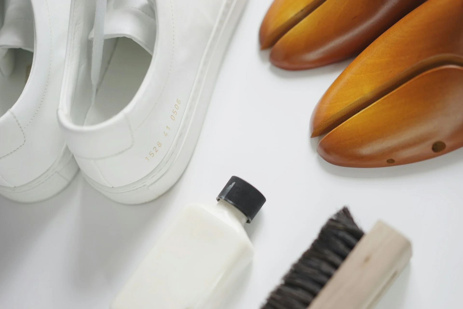 Sepasang sneaker putih di samping sikat sepatu, botol cairan pembersih, dan shoe tree kayu