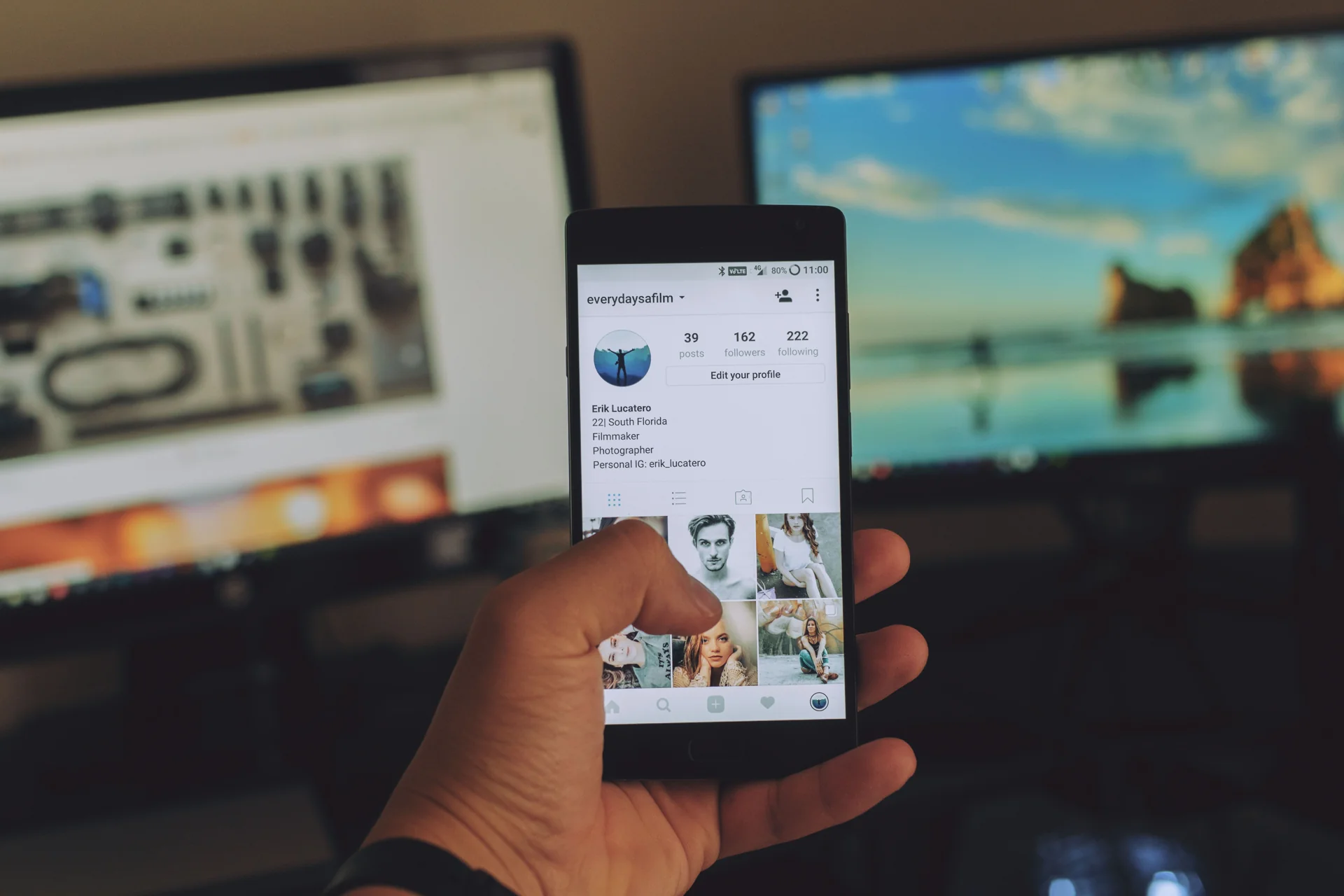 Tangan memegang HP yang menampilkan halaman profil Instagram di depan dua monitor