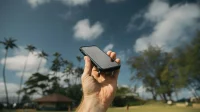Tangan memegang smartphone di luar ruangan saat siang terik dengan langit biru dan pohon kelapa