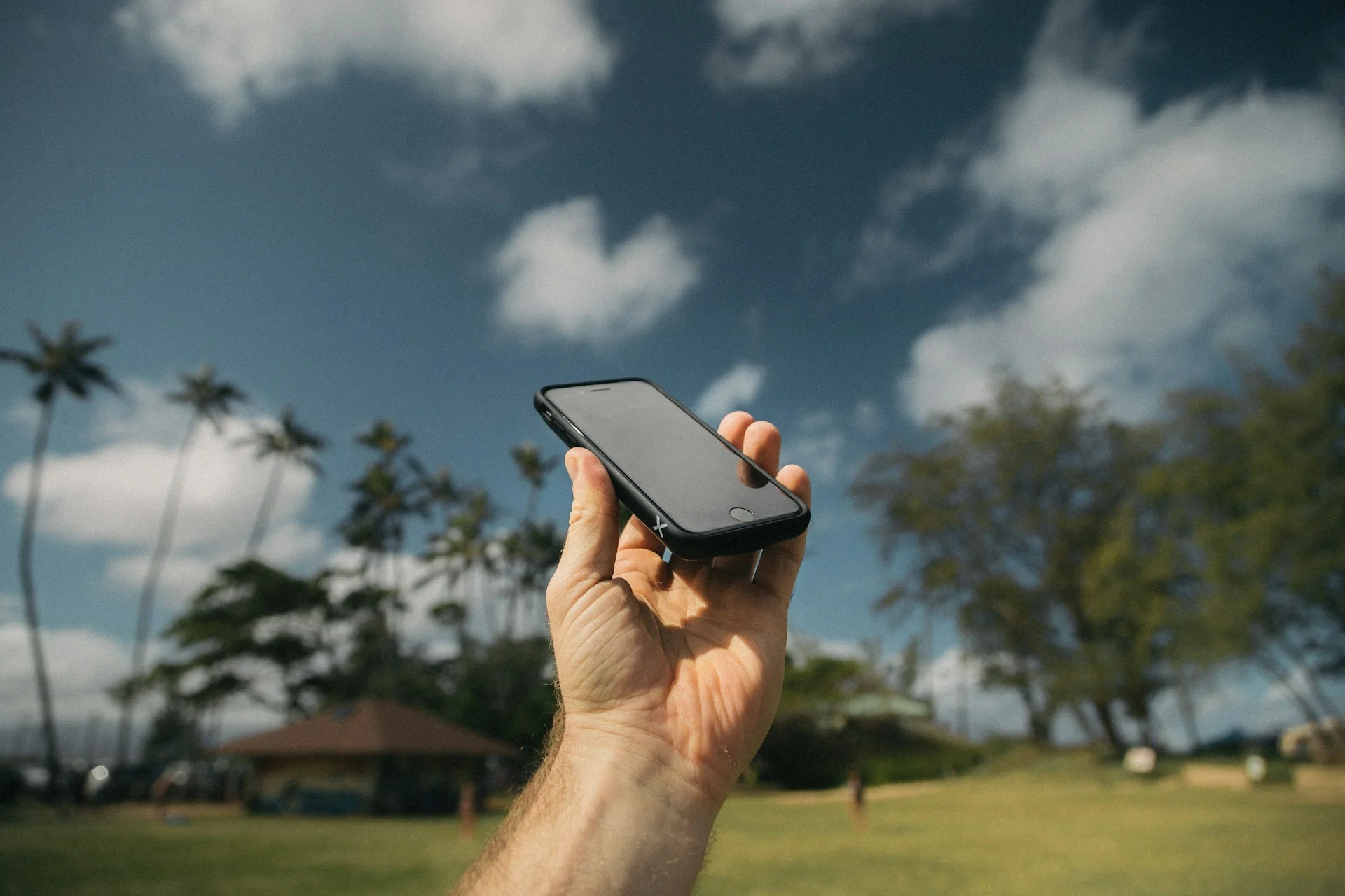 Tangan memegang smartphone di luar ruangan saat siang terik dengan langit biru dan pohon kelapa