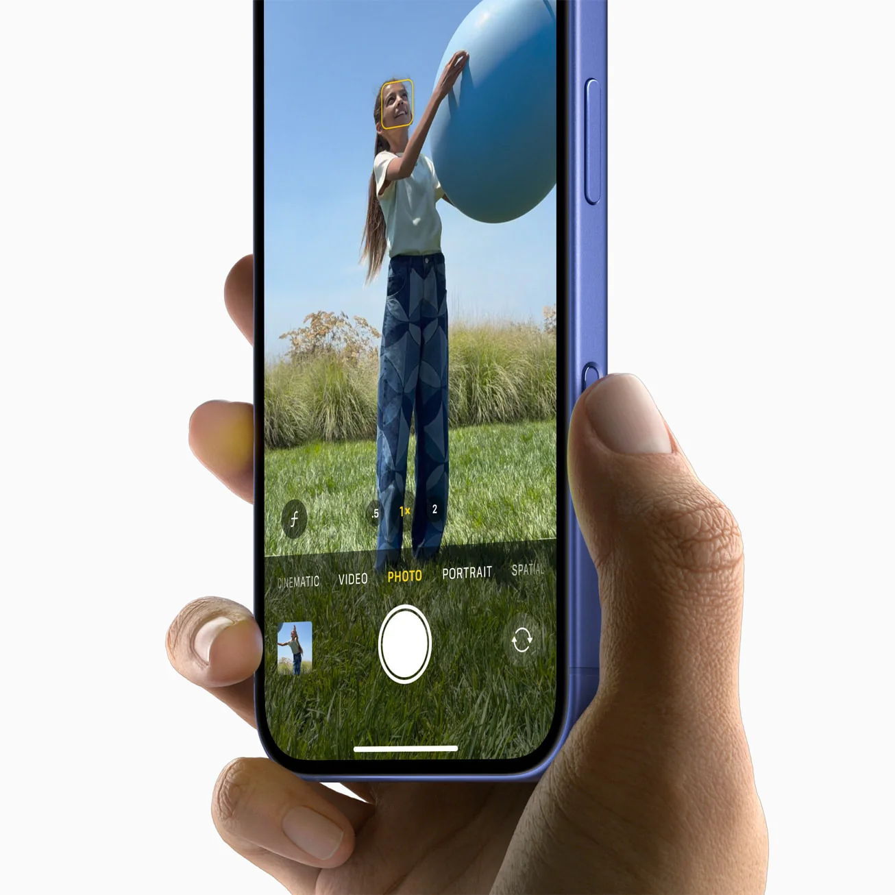 Tangan memegang iPhone 16 biru dengan jempol di tombol Camera Control saat aplikasi kamera terbuka
