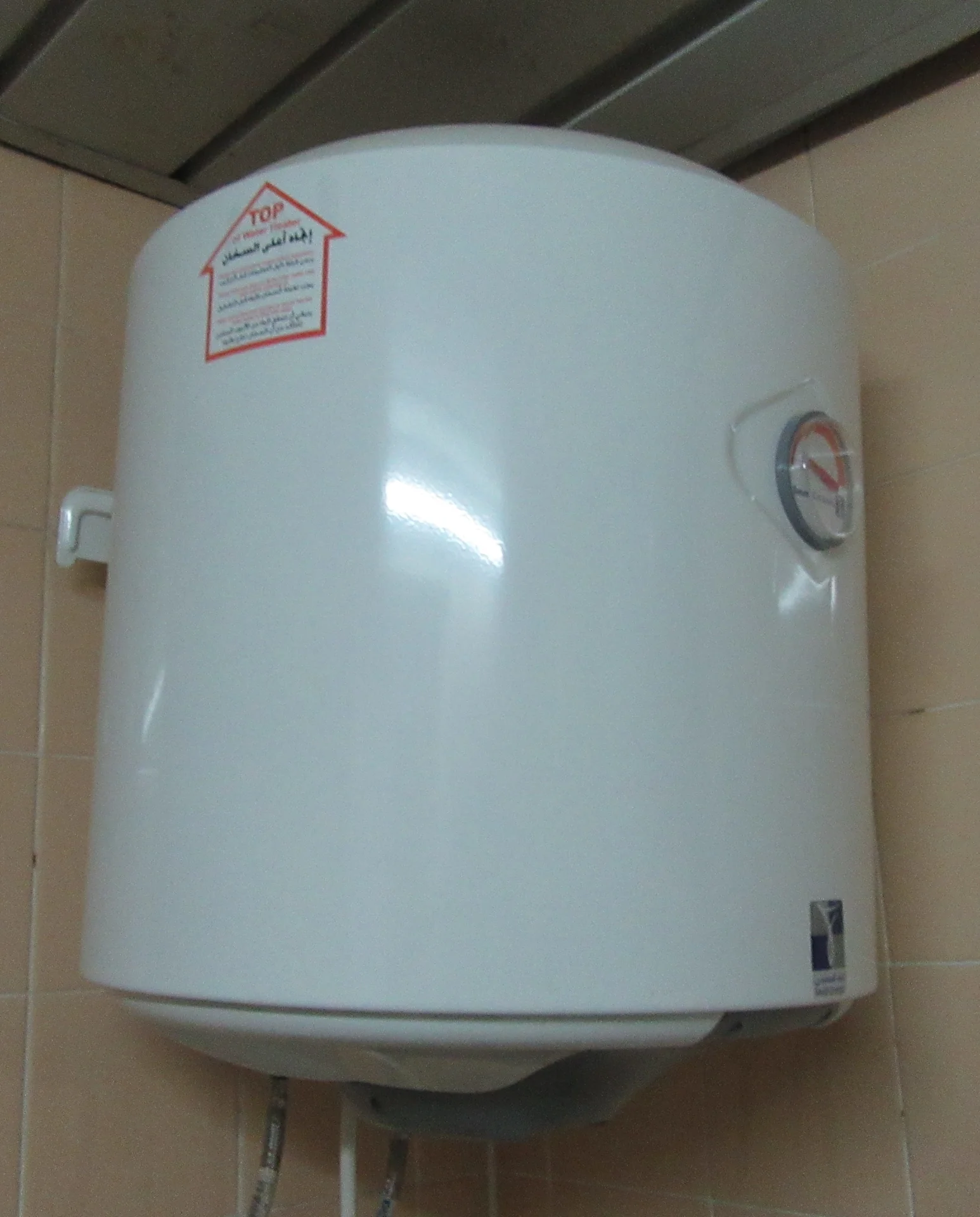 Water heater listrik tipe tangki berwarna putih terpasang di dinding keramik dekat plafon, dengan indikator suhu bundar